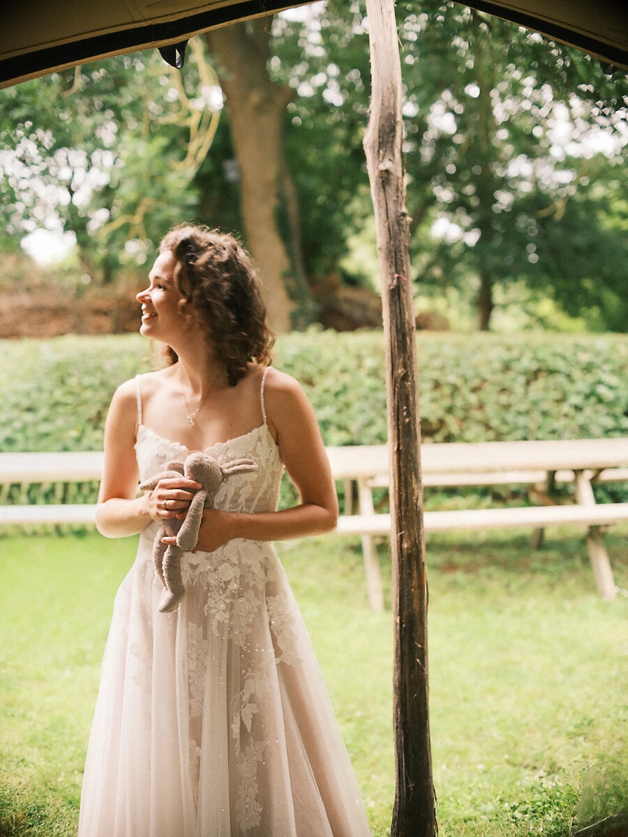 Wedding SP23 Laughing under the edge of a marquee in a strappy ivory gown, the bride holds a soft toy rabbit, a playful wedding portrait