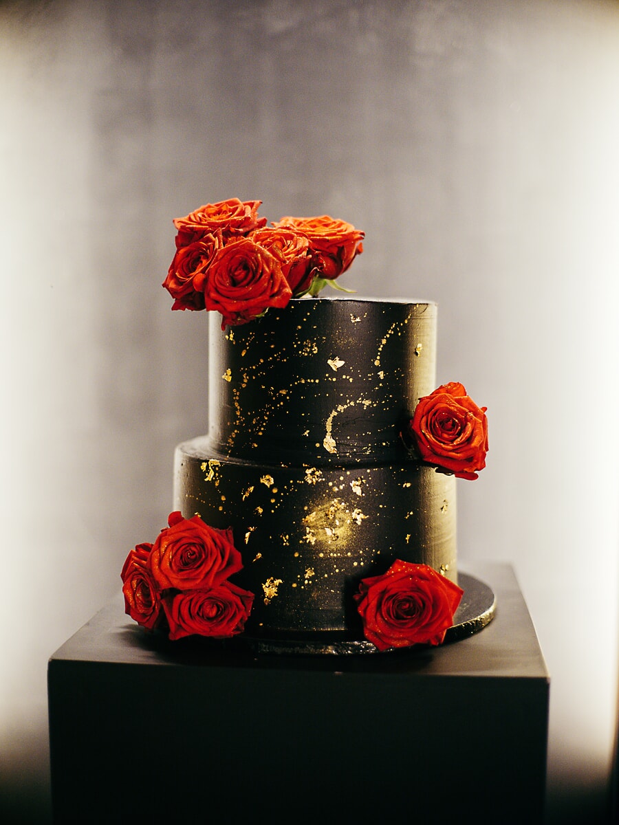 Wedding SP24 Two-tier black and gold cake topped with red roses on a dark plinth, a striking detail from the wedding photography