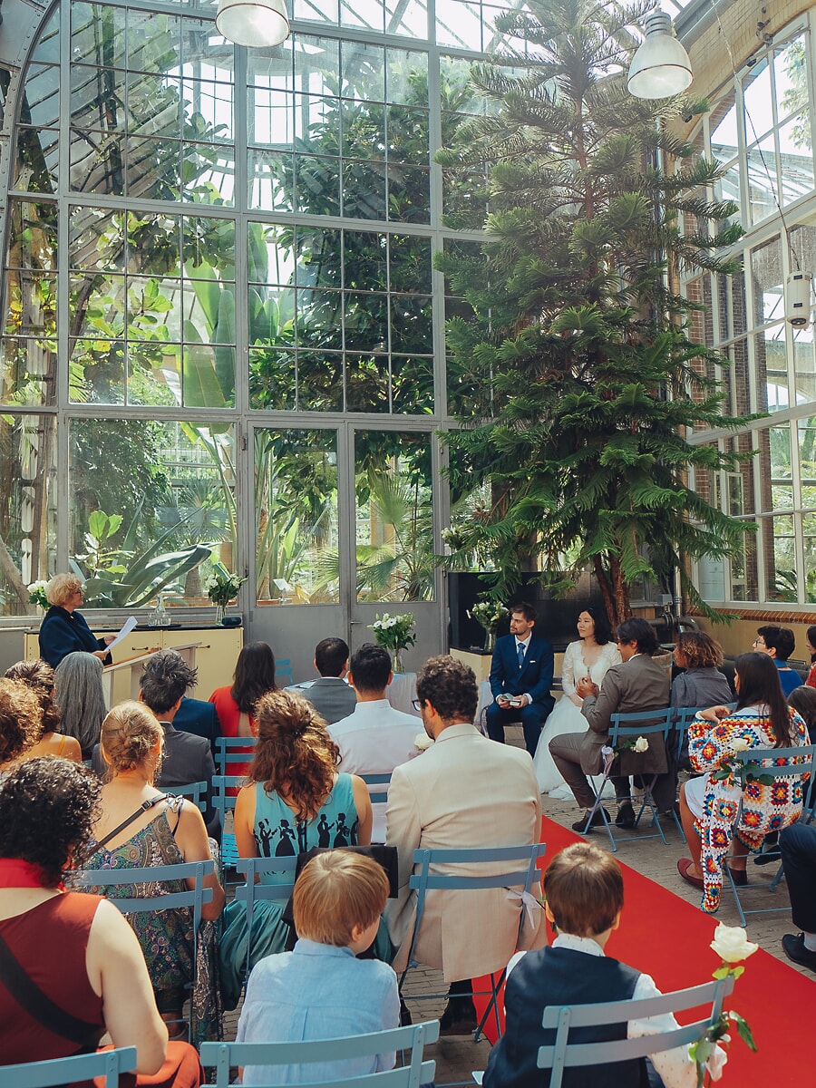 Wedding SP25 Guests on blue chairs watch the ceremony in a glass orangery, a red runner leading to the couple, by a wedding photographer in Amsterdam