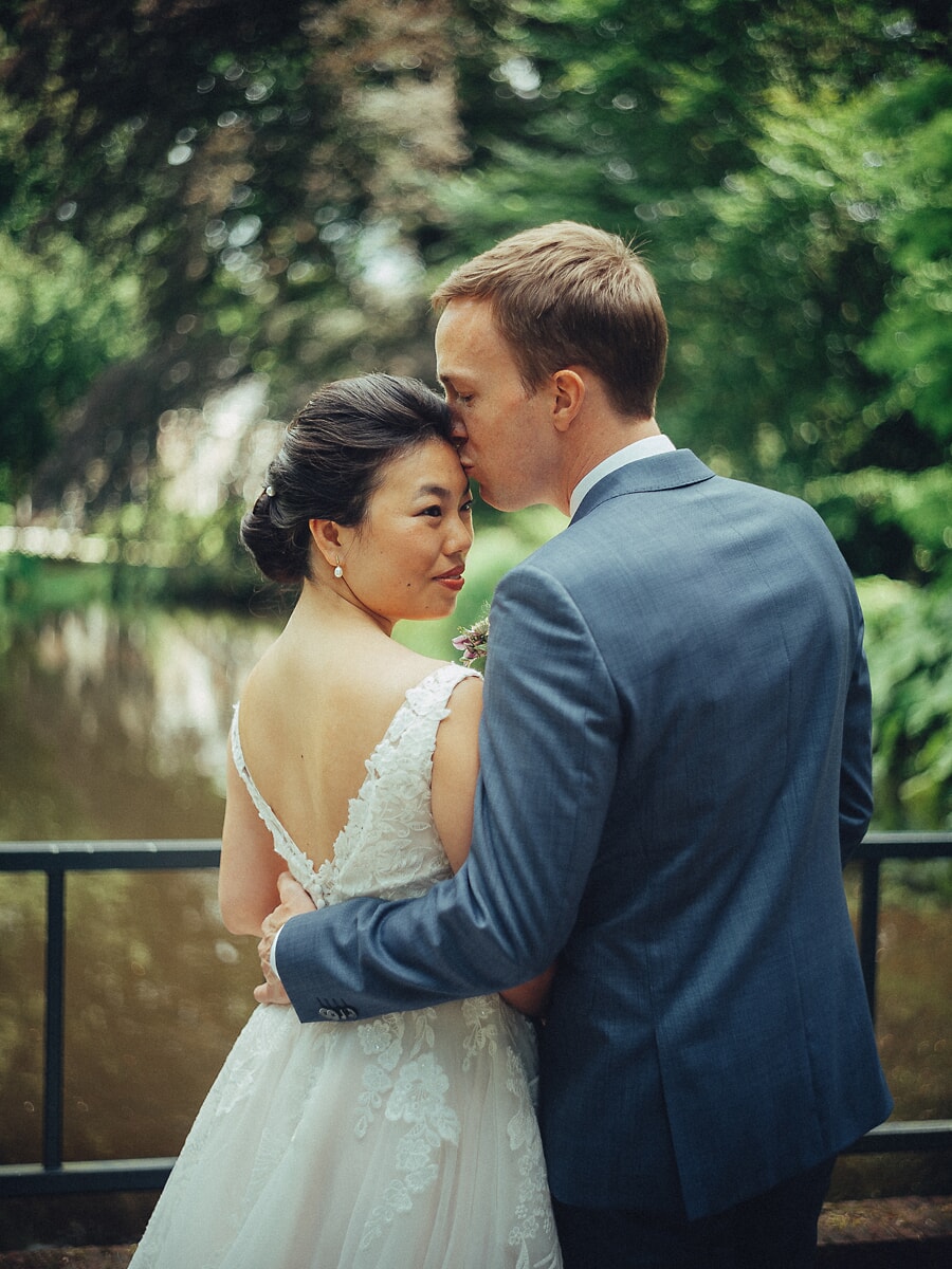 Wedding SP26 Groom in a blue suit kisses his bride's forehead beside a green canal railing, her lace back gown catching the light, a tender newlywed portrait