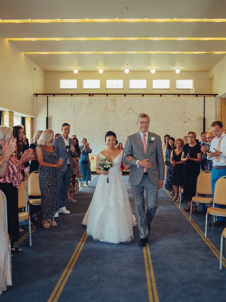 Wedding SP31 Arm in arm with her father, the bride walks the blue aisle as guests applaud from both sides, a documentary ceremony photograph