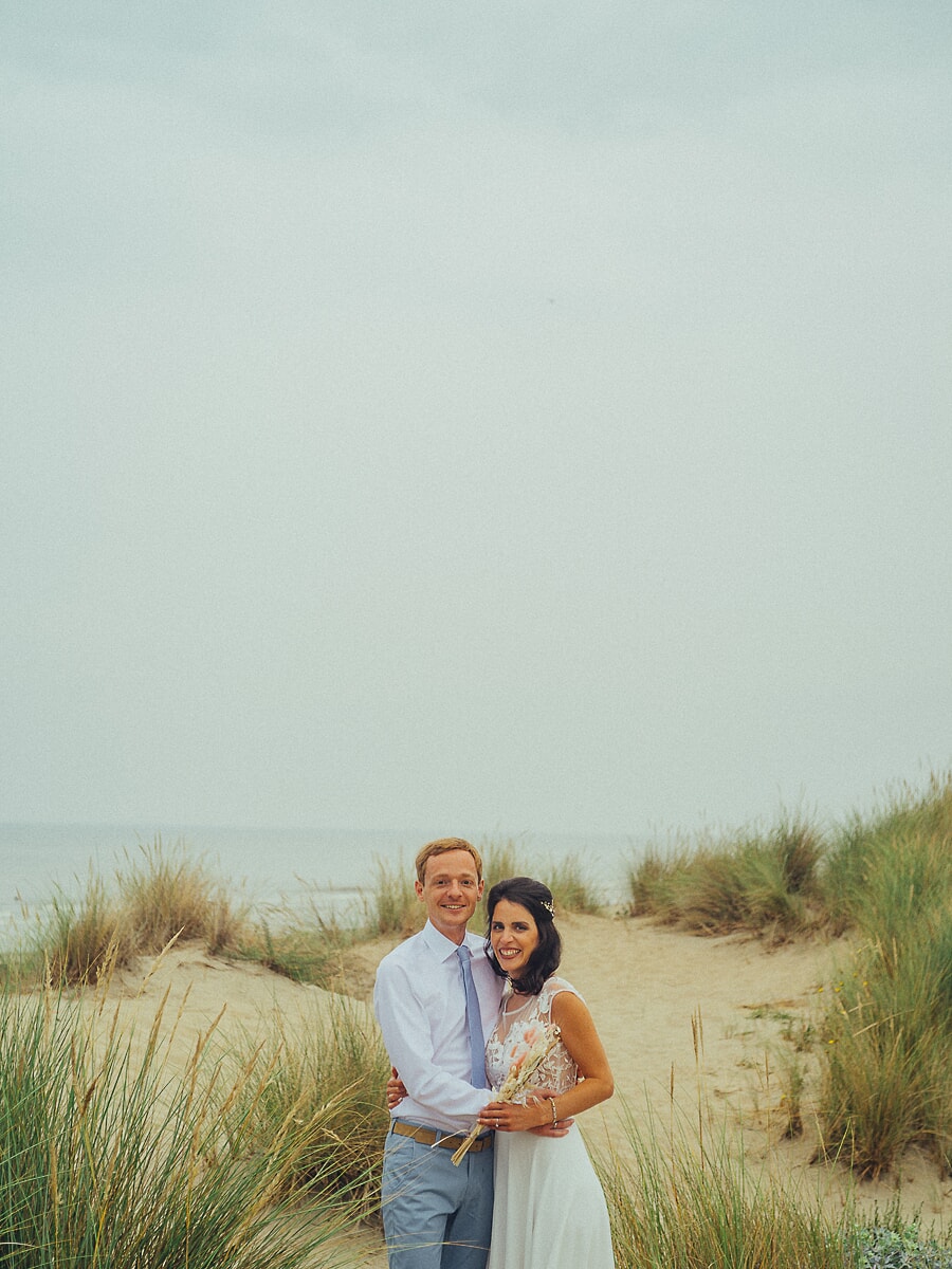 Wedding SP33 Couple embrace among the dune grass in soft coastal light, the bride holding a dried flower bouquet, a relaxed beach wedding portrait
