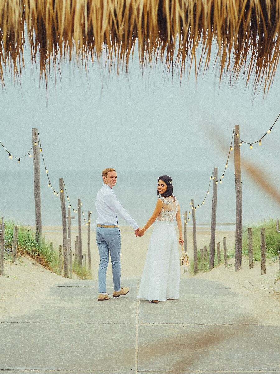 Wedding SP34 Under strings of lights, the newlyweds glance back while walking toward the dunes, photographed by a wedding photographer in Amsterdam