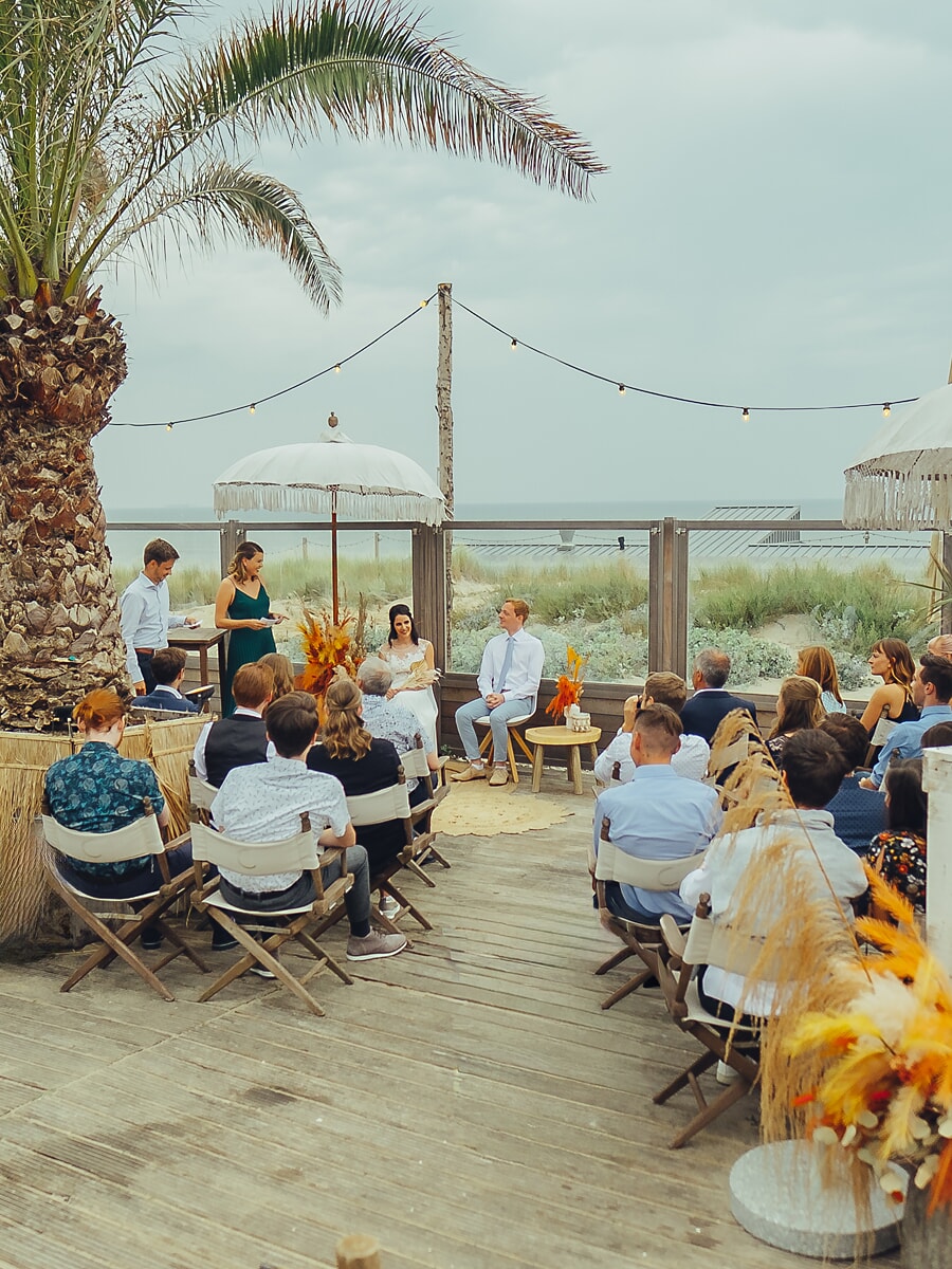 Wedding SP36 Guests on a wooden beach deck face the seated couple beneath a fringed parasol, captured by a wedding photographer in Amsterdam