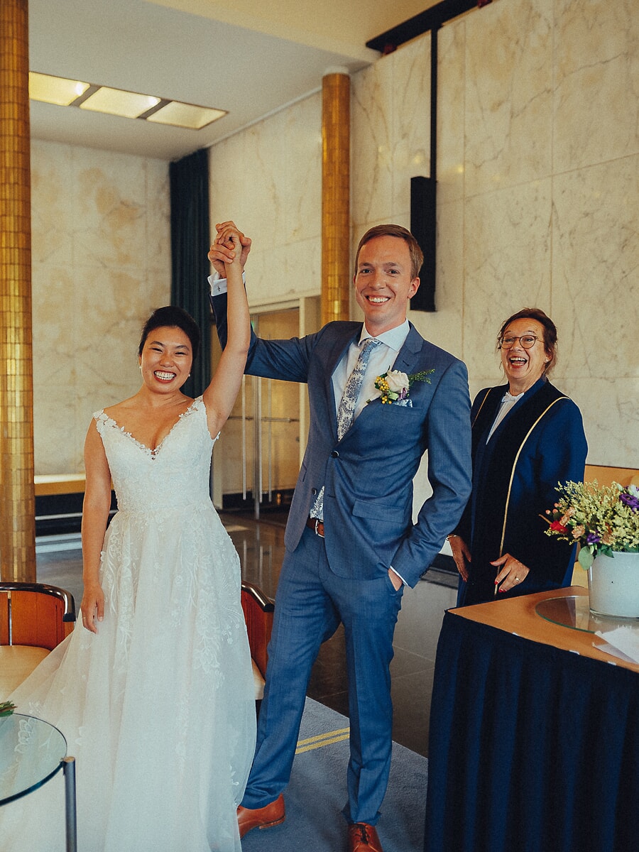 Wedding SP37 Laughing groom lifts his bride's hand in triumph beside the registrar in a marble city hall, a joyful ceremony photo