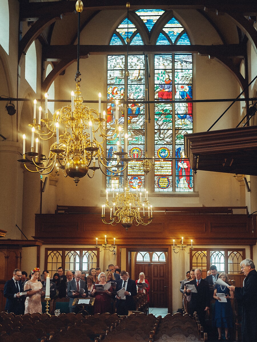 Wedding SP38 Beneath stained glass and brass chandeliers, guests stand with hymn sheets, a timeless frame by a wedding photographer in Amsterdam