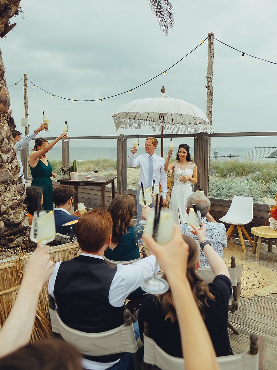 Wedding SP39 Raising their glasses toward the couple under a parasol at a beachside venue, guests share a festive toast, a lively wedding photograph