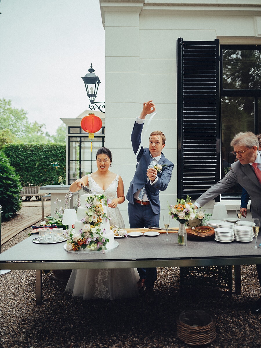 Wedding SP41 Groom raises the knife overhead as his bride cuts the floral tiered cake on the terrace, a playful reception photo