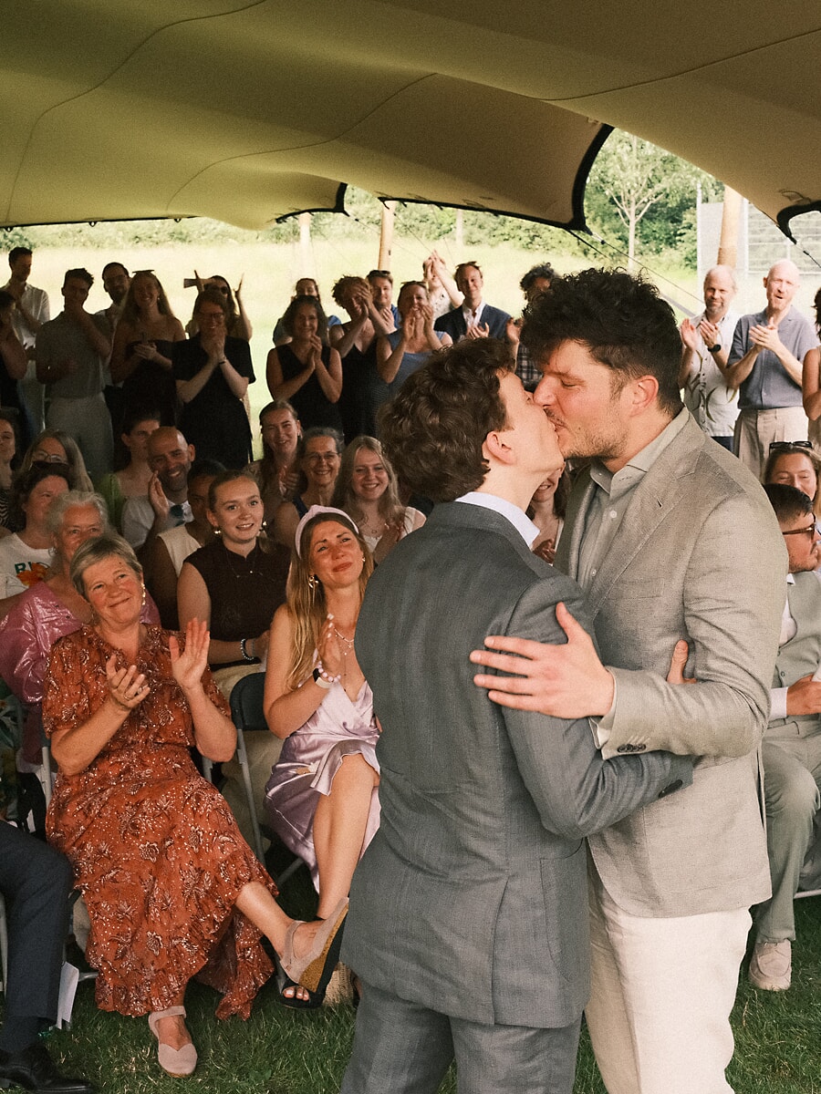 Wedding SP8 Two grooms share their first kiss under a marquee while seated guests applaud, a candid same-sex wedding photograph