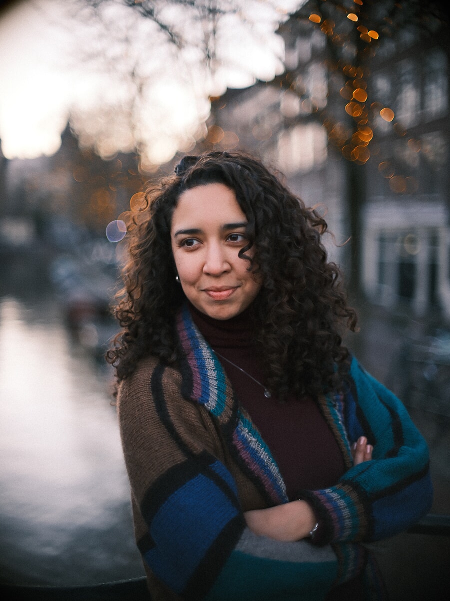 Woman with dark curls and folded arms in a striped knit cardigan, string lights glowing behind her, a confidence portrait at dusk in Amsterdam