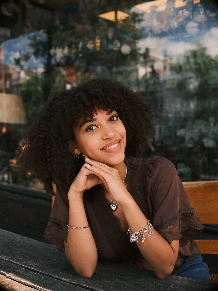 Woman resting her chin on her hands at a cafe table before a painted canal scene, a lifestyle portrait in an Amsterdam cafe