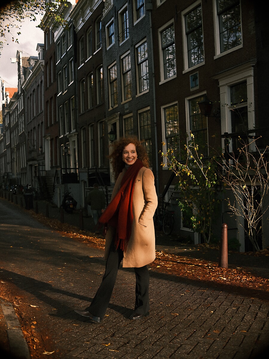 Woman in a camel coat and red scarf standing in an autumn canal street strewn with leaves, an autumn portrait in Amsterdam