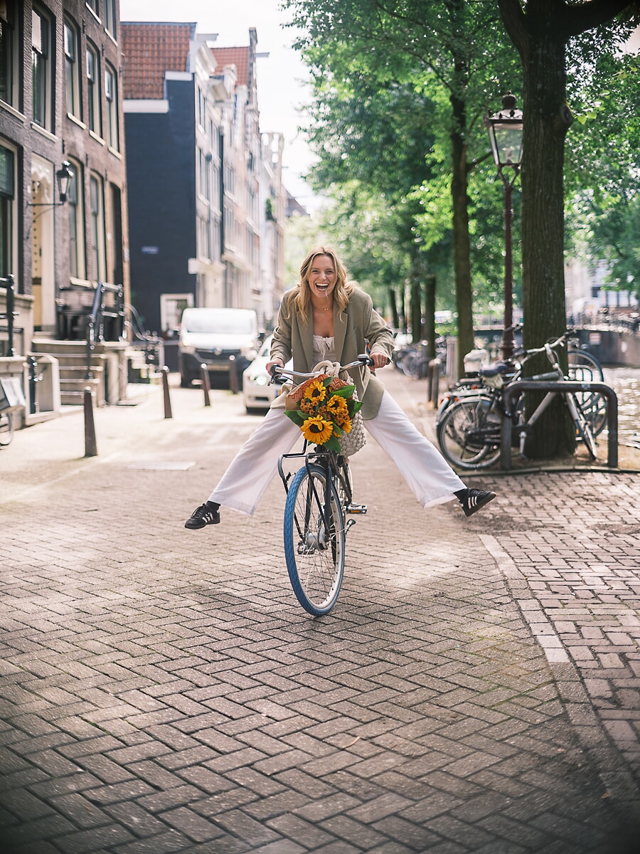 Woman freewheeling toward the camera with her legs kicked out, laughing, sunflowers on the handlebars, an Amsterdam street portrait