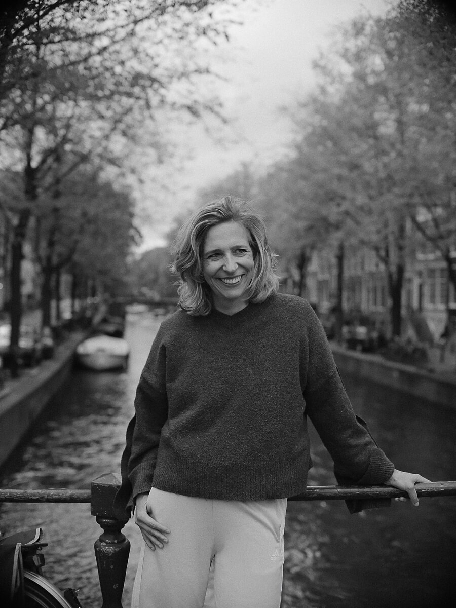 Blonde woman laughing as she leans back on a canal railing, a black-and-white confidence portrait among autumn trees