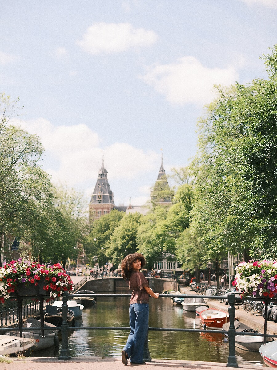Woman with curly hair looking back from a bridge railing with the Rijksmuseum towers behind, portrait photography in Amsterdam