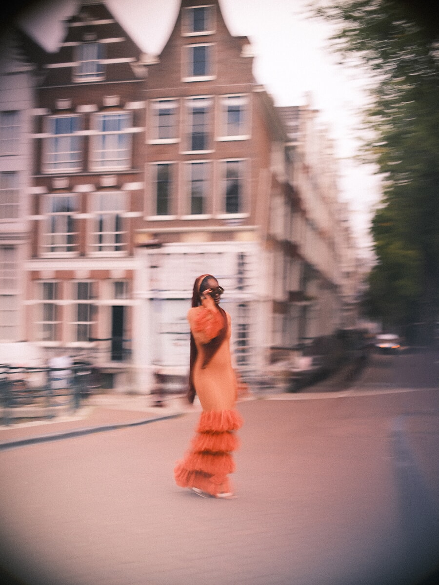Woman in an orange ruffled dress crossing the street in a panning blur, the gabled houses streaked behind her