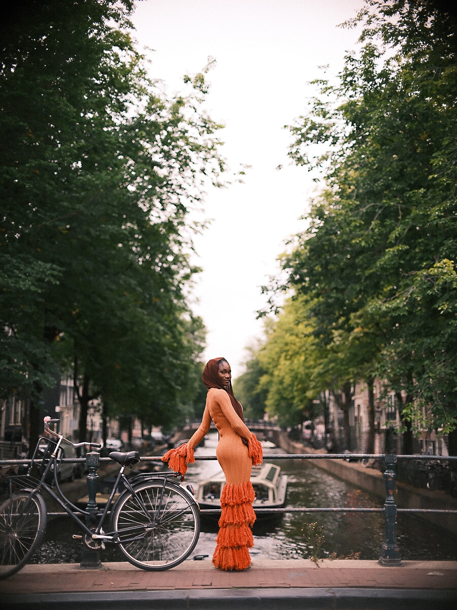 Woman in an orange fringed knit dress and head wrap glancing back on a bridge, a colourful portrait in Amsterdam