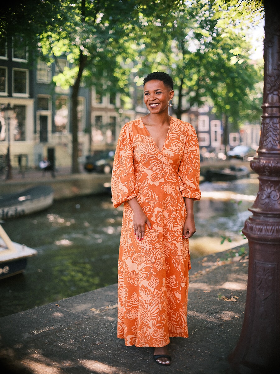 Woman in an orange leaf-print maxi dress beside a canal with an ornate lamppost, a full-length confidence portrait in Amsterdam