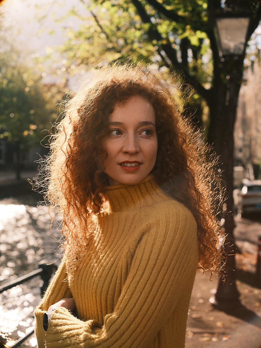Woman with red curls in a mustard turtleneck glancing over her shoulder in golden autumn light, portrait photography in Amsterdam