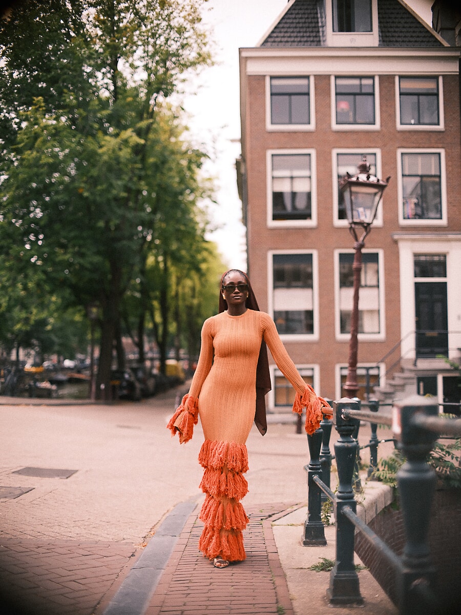 Woman in an orange fringed dress walking beside a canal railing below a brick gabled house, a street portrait in Amsterdam