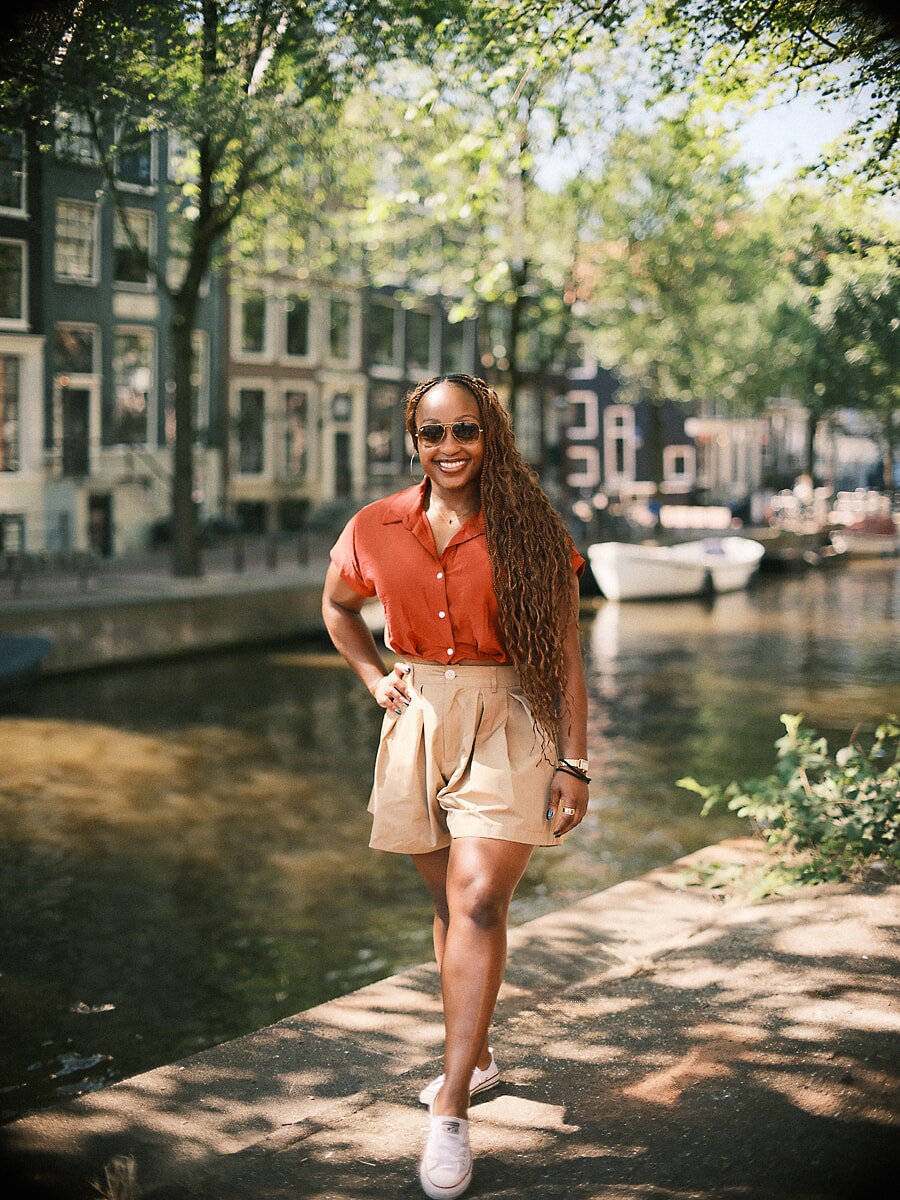 Woman in a rust shirt and sunglasses walking a sunlit canal quay during her portrait photoshoot in Amsterdam