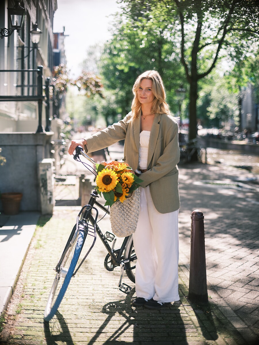 Woman in an oversized blazer and white trousers standing with a bicycle, sunflowers in a net bag on the handlebars, a portrait in Amsterdam