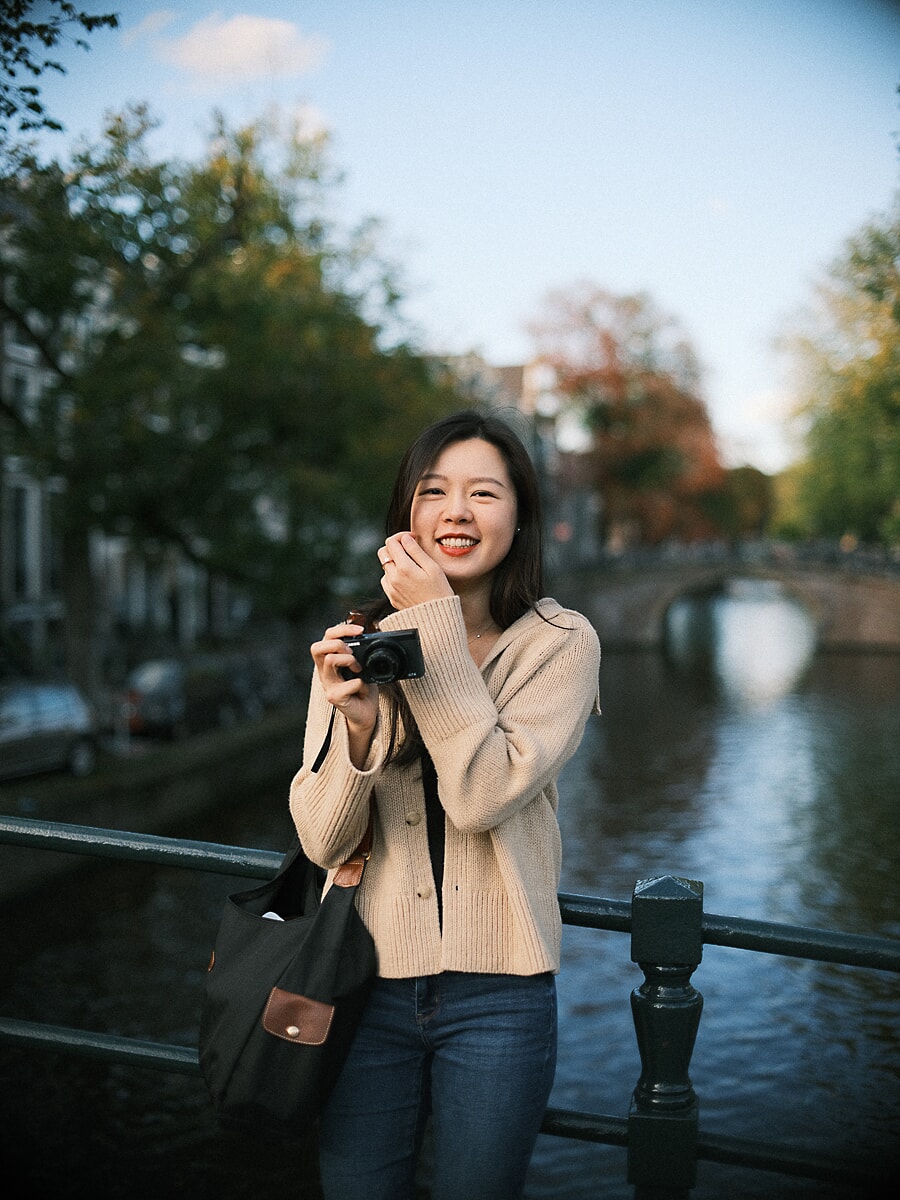 Woman in a cream cardigan laughing with a small camera in her hands on a canal bridge, a warm portrait from her dating photoshoot in Amsterdam
