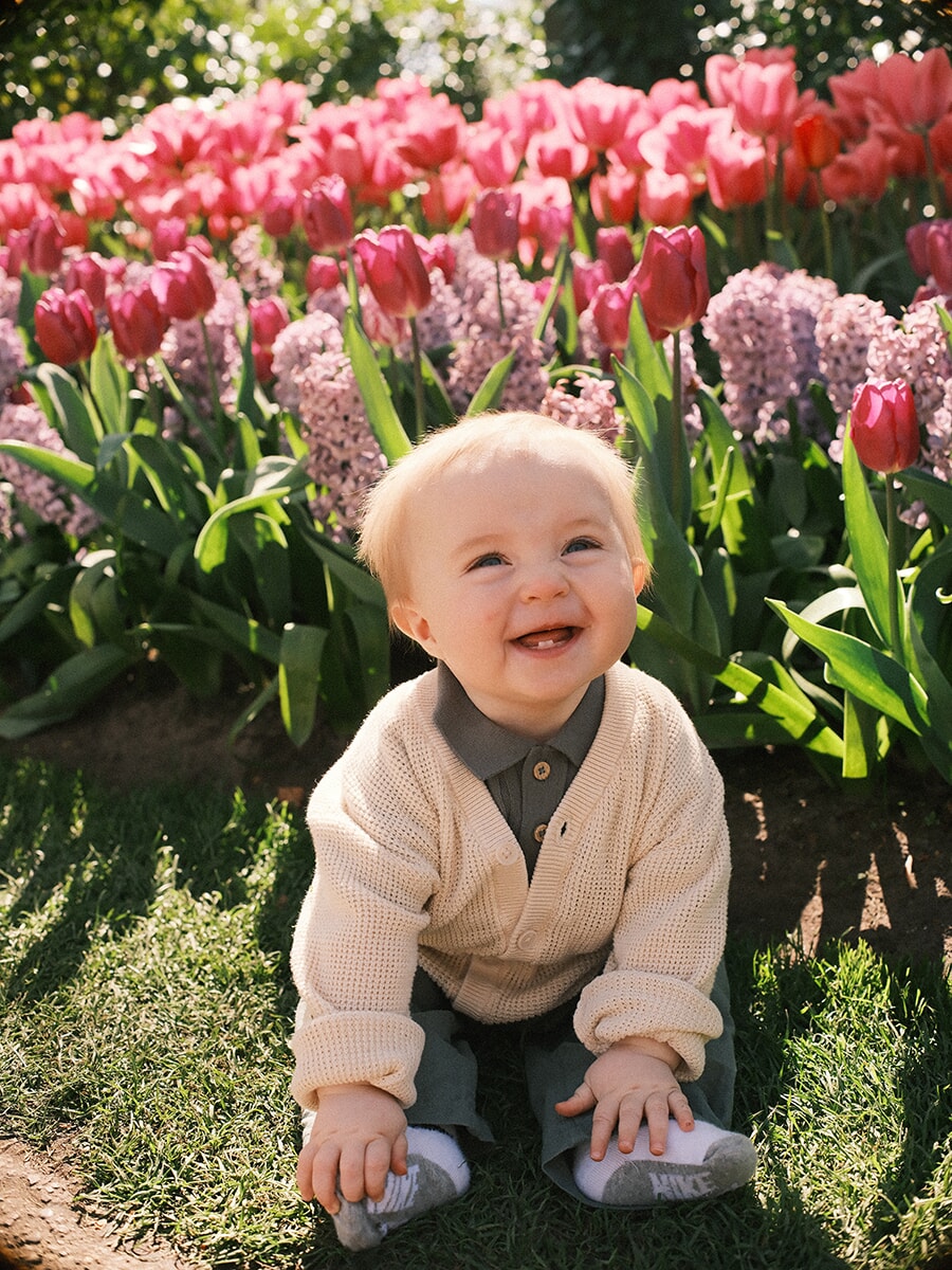Laughing baby in a cream knit cardigan sitting on the grass, pink tulips and lilac hyacinths behind