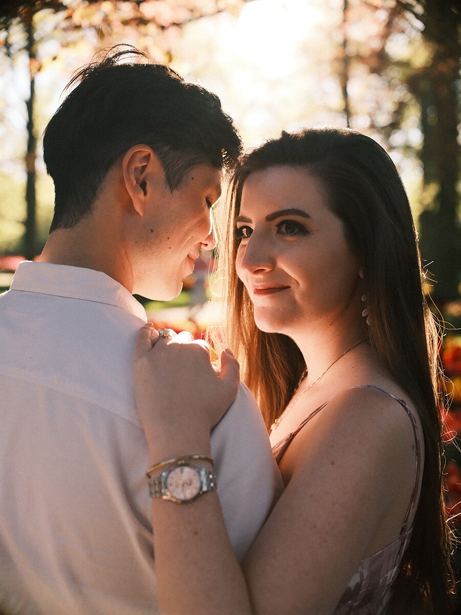 Backlit couple with faces almost touching, her ring hand resting on his shoulder in warm golden light