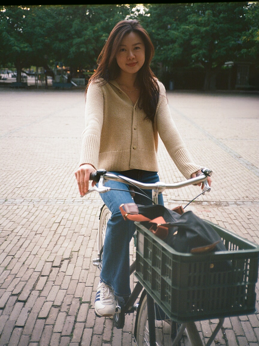 A woman in a cream cardigan sits on a bicycle with a front crate for an analog portrait in Amsterdam
