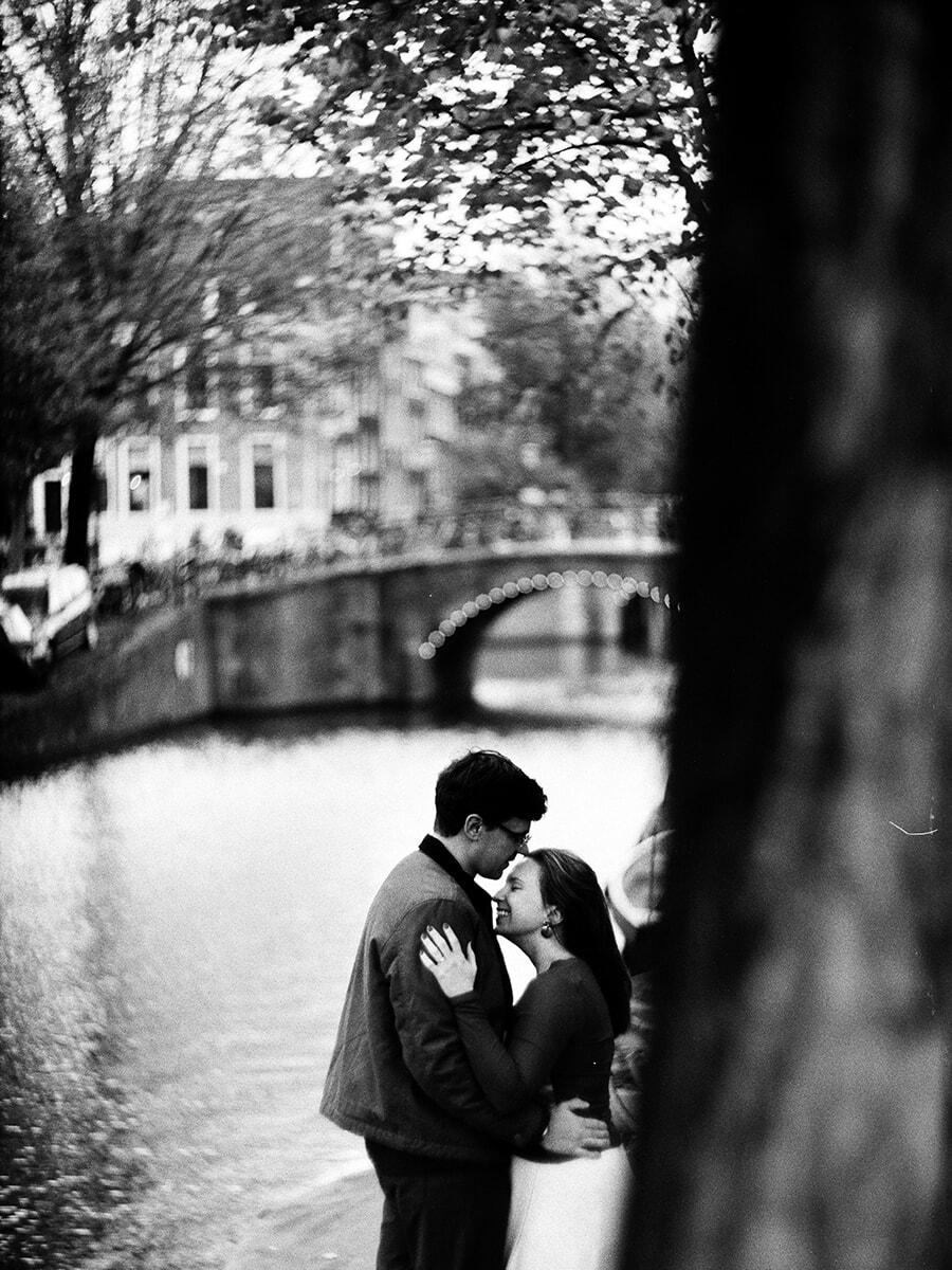 A forehead kiss beside the canal, with a blurred tree trunk in the foreground and an arched bridge behind the couple