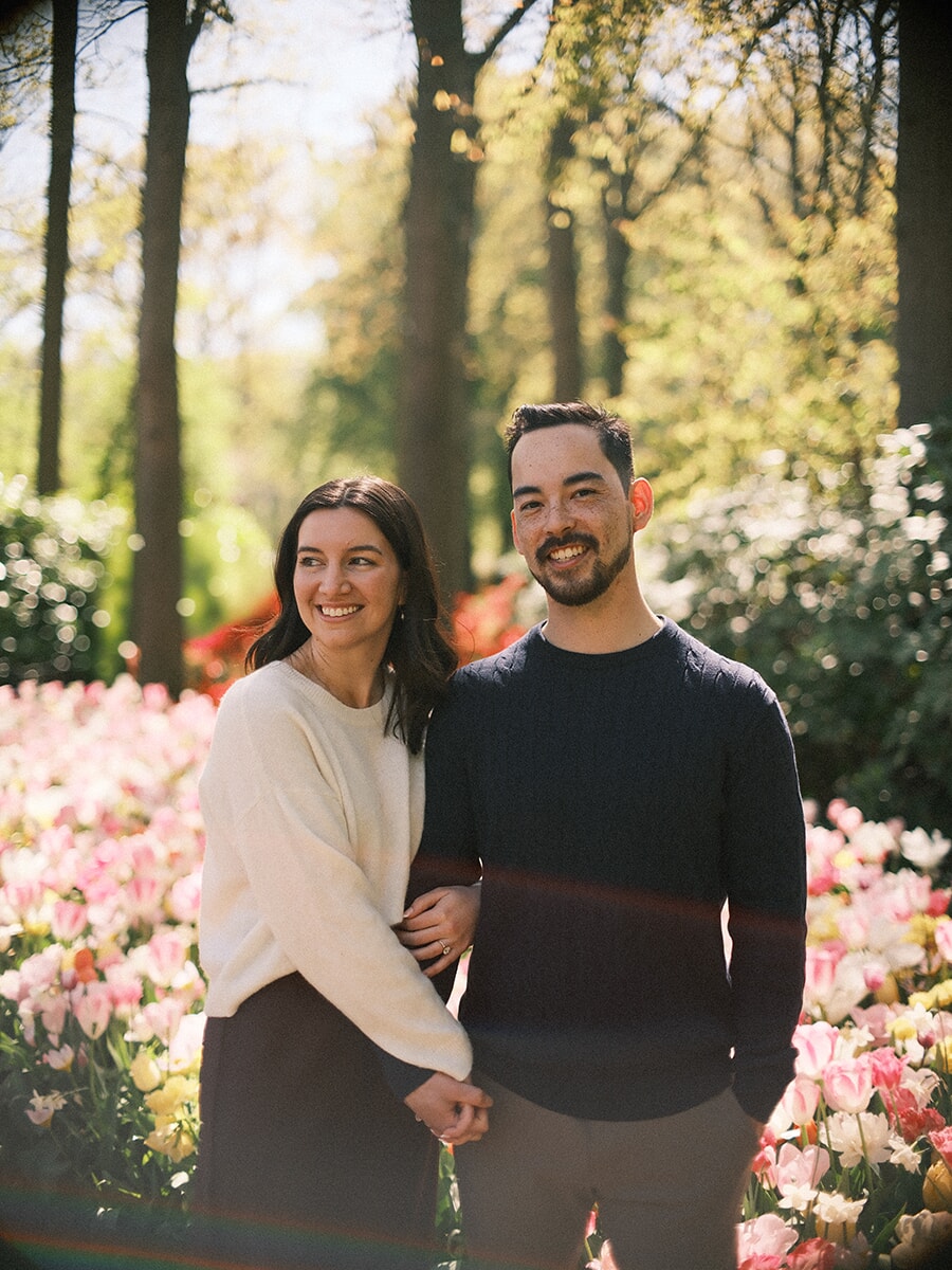 Smiling couple arm in arm among pale pink and white tulips at Keukenhof, sun flare streaking across the frame
