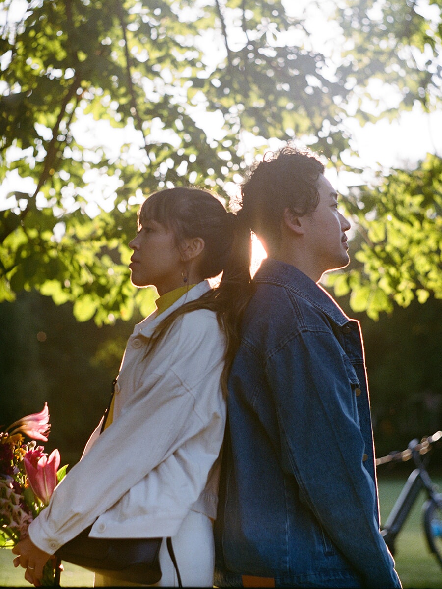Back to back beneath sunlit leaves, a woman in a white jacket holds pink flowers beside her partner in a denim jacket