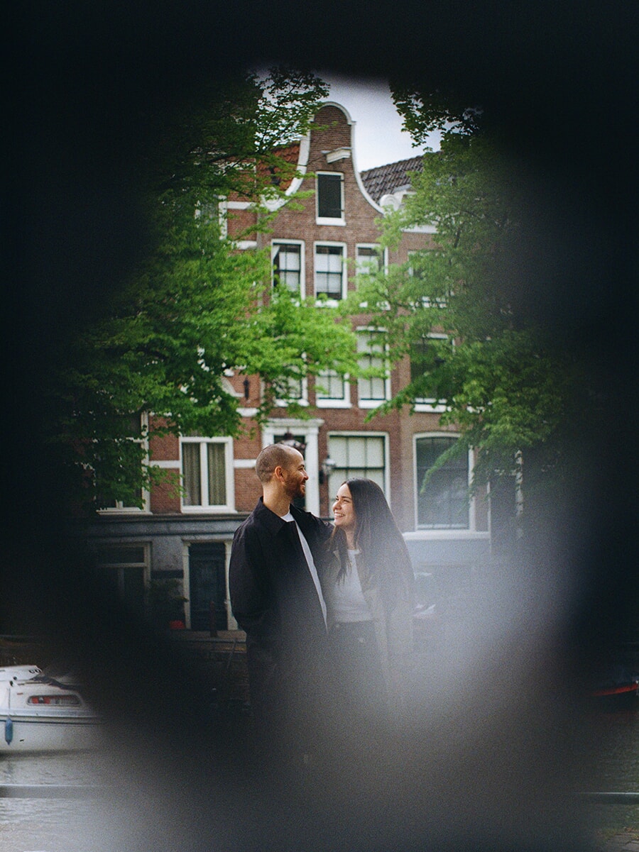 Through a dark, blurred foreground, a couple smile at each other beside an Amsterdam canal in a colour film portrait