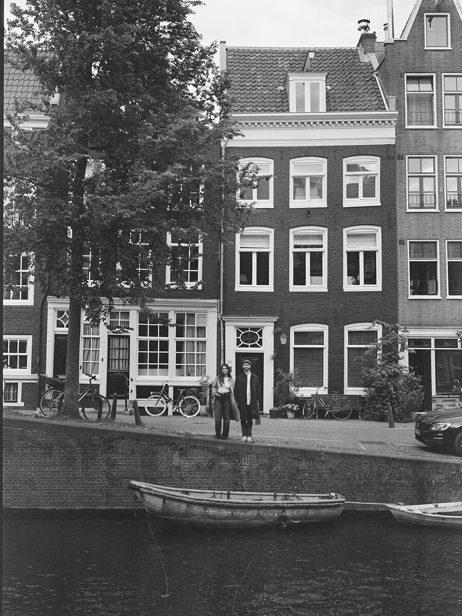 Analog couple portrait across an Amsterdam canal, with tall brick houses behind the pair and a moored rowing boat below