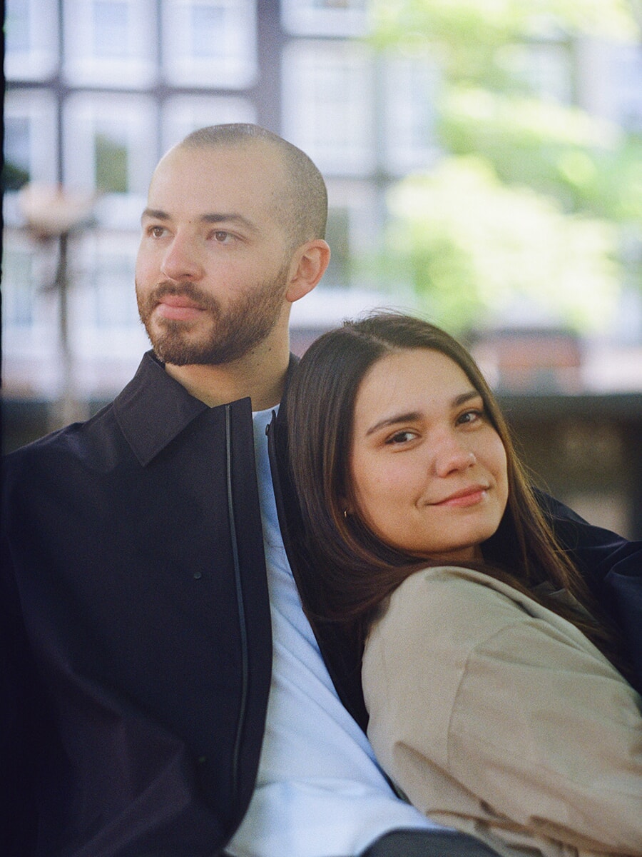 Leaning against her partner in a dark coat, a woman in beige smiles at the camera in a colour film portrait