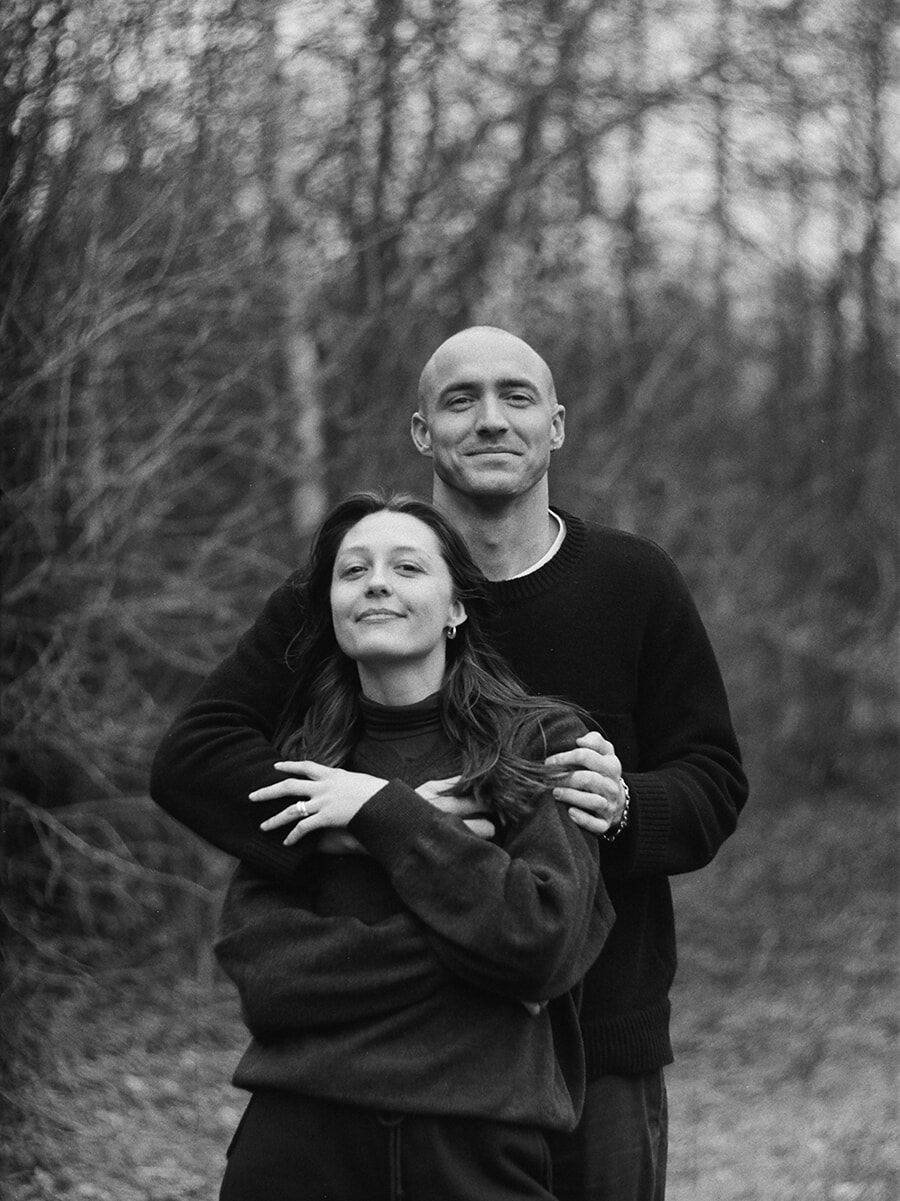 With his arms around his partner from behind, a man faces the camera with her among the trees