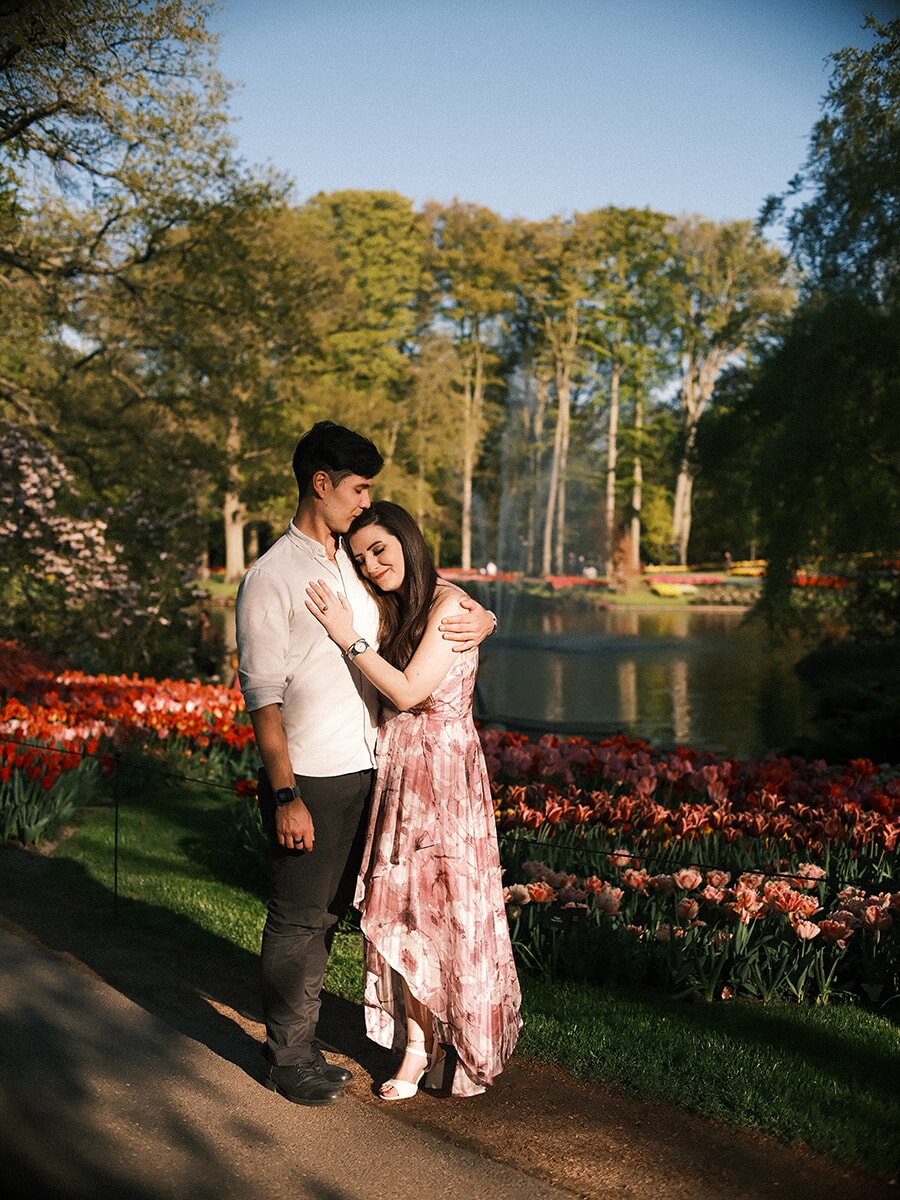 Eyes closed in an embrace on a sunlit path, a couple with a fountain pond and red tulip beds glowing behind them