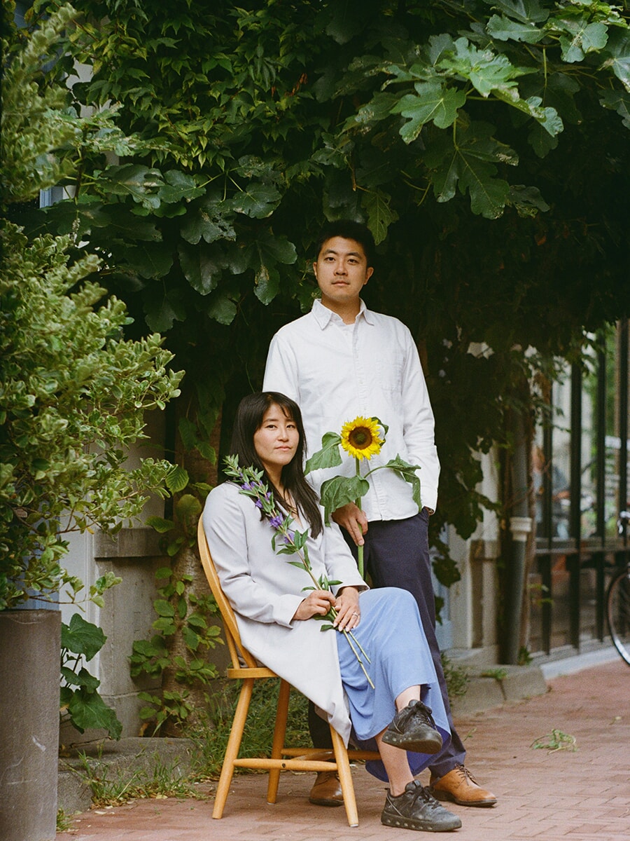 Among dense green leaves, a woman sits holding purple flowers beside her standing partner with a sunflower