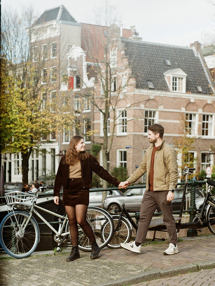 Hand in hand across a canal bridge, a woman in a brown skirt and a man in a tan jacket walk past parked bicycles