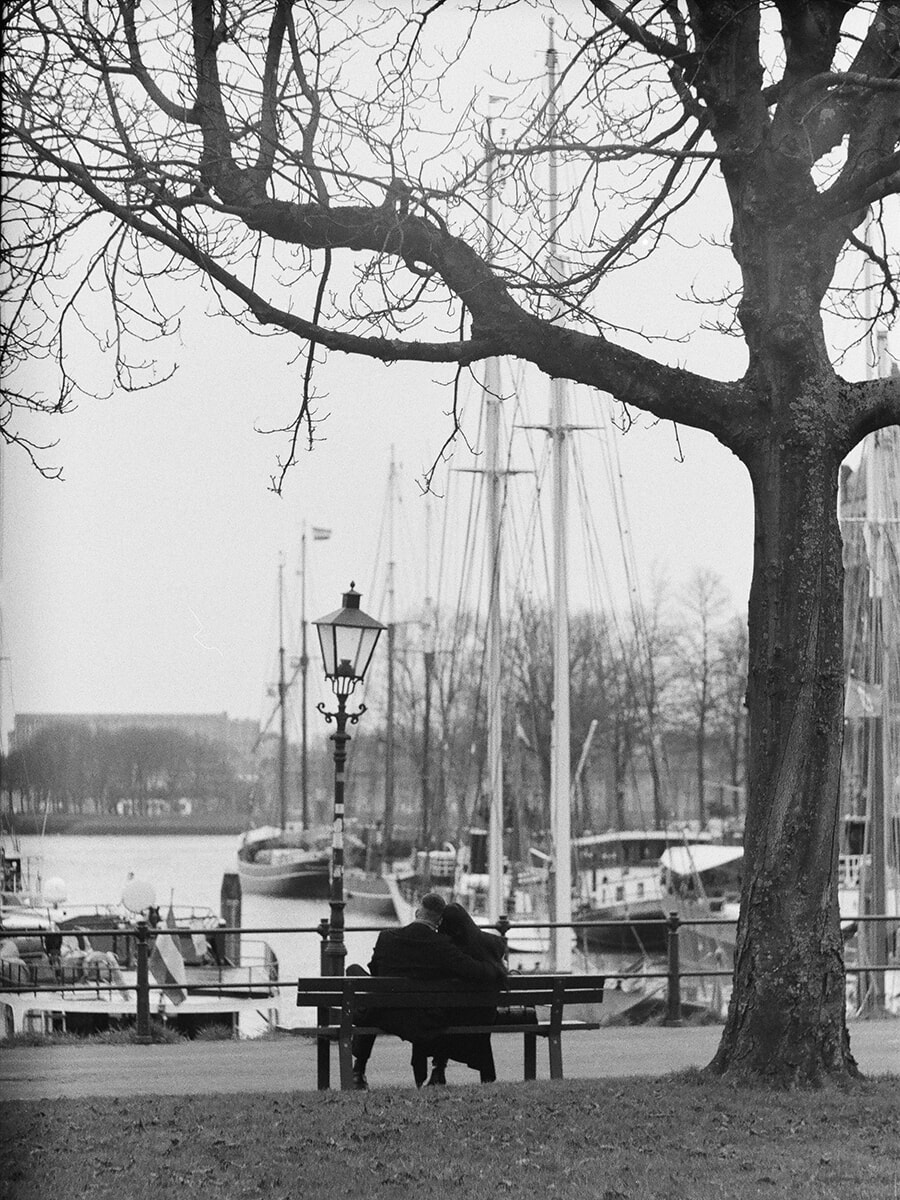 Seen from behind, a couple lean close on a bench beneath bare branches, overlooking moored sailing boats