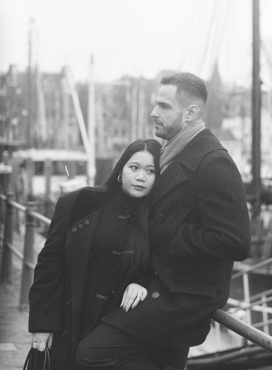 At a harbour railing, a woman rests her head against her partner’s chest, with sailing-boat masts behind them