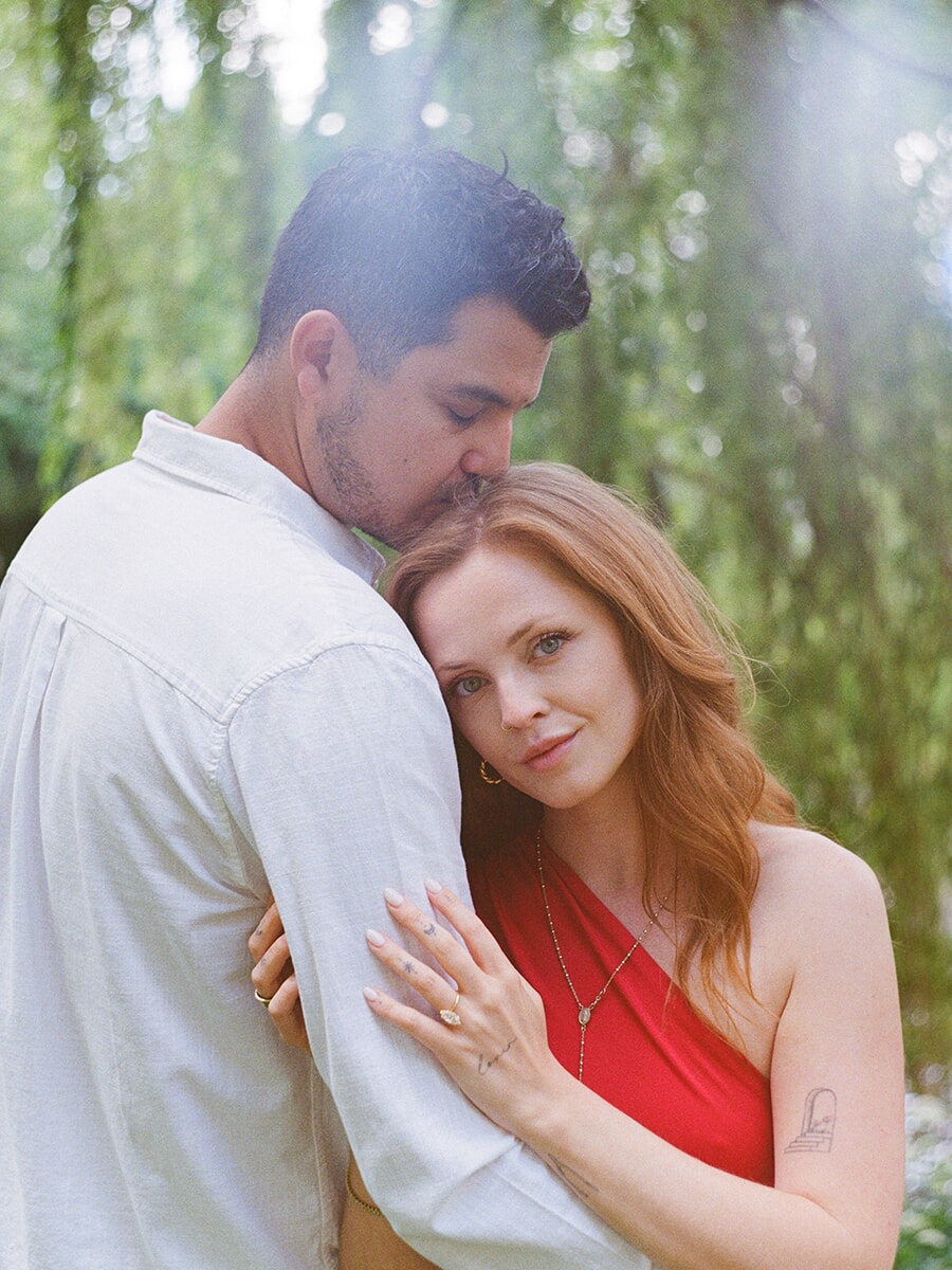 In a colour film close-up, a woman in a red dress looks at the camera as her partner kisses her head