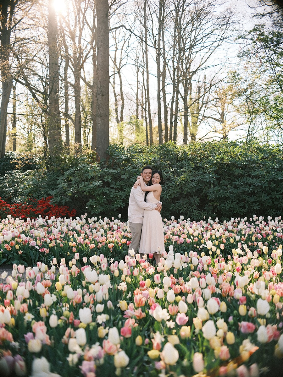 Couple hugging in a tulip field of white, pink and cream blooms, sunlight breaking through the trees behind them