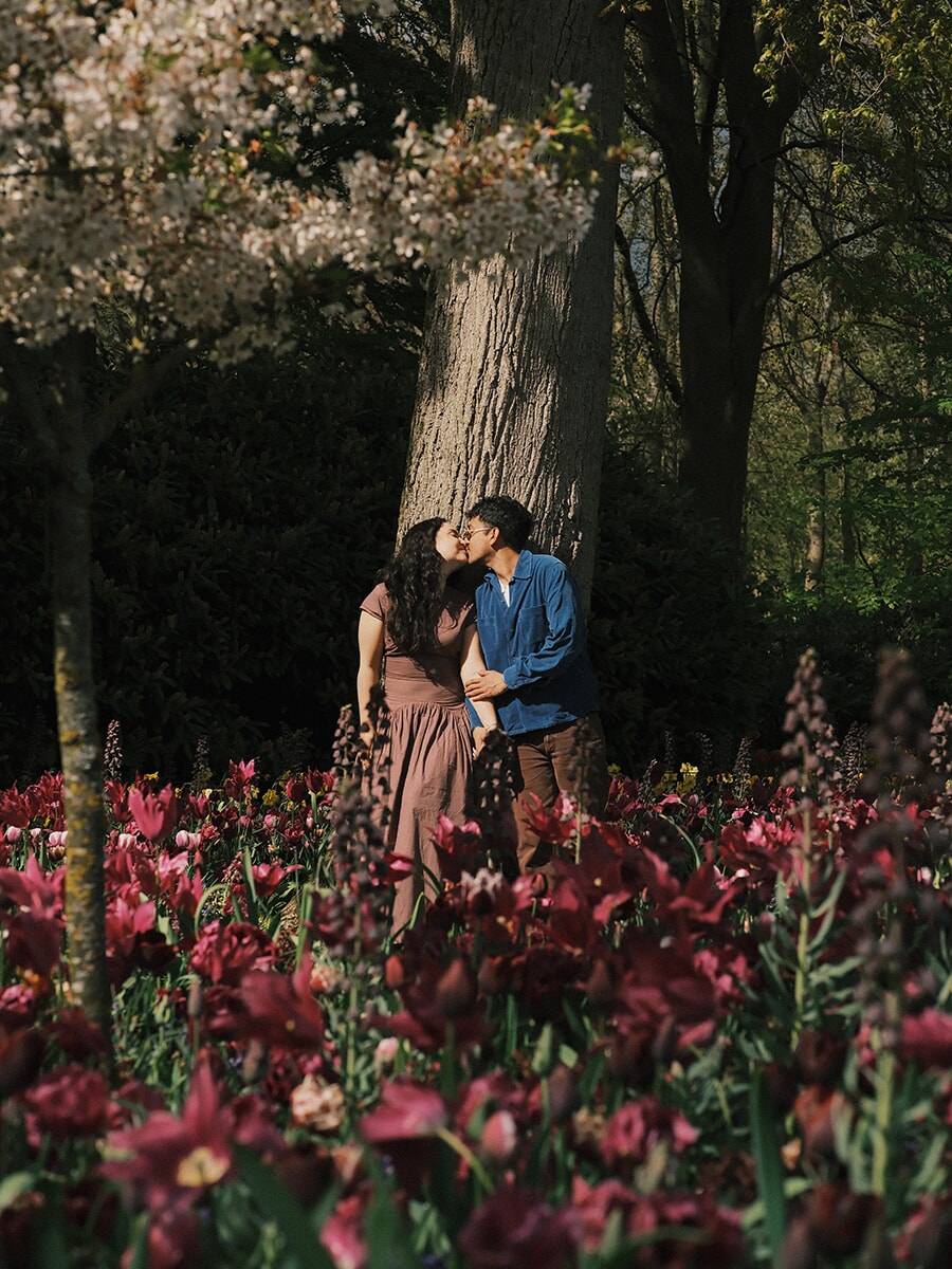 Couple kissing against a big tree trunk under white blossom, burgundy tulips and tall dark flower spikes in front