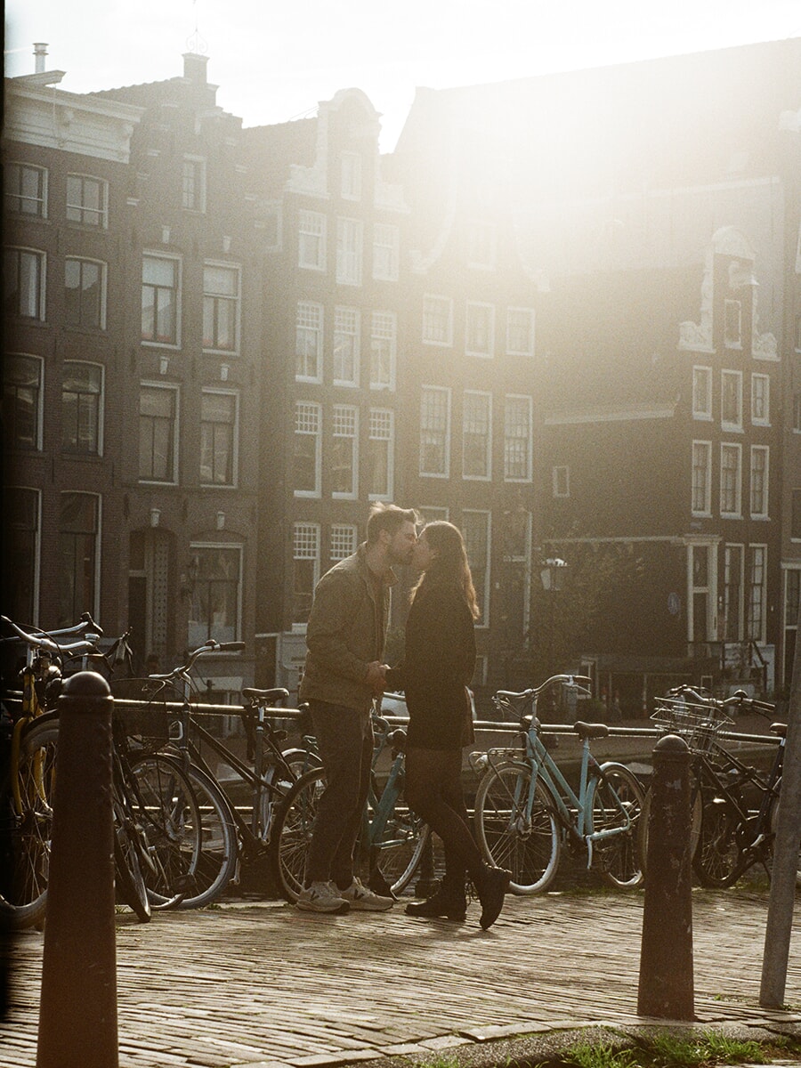 An analog couple photoshoot on an Amsterdam canal bridge, with a kiss among parked bicycles and backlit gabled houses behind