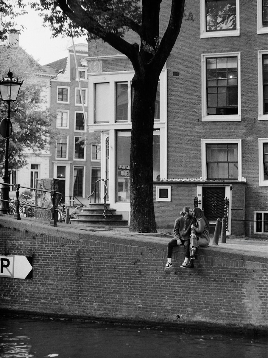 Beneath a tree on an Amsterdam canal wall, a seated couple kiss in a wide black-and-white film portrait