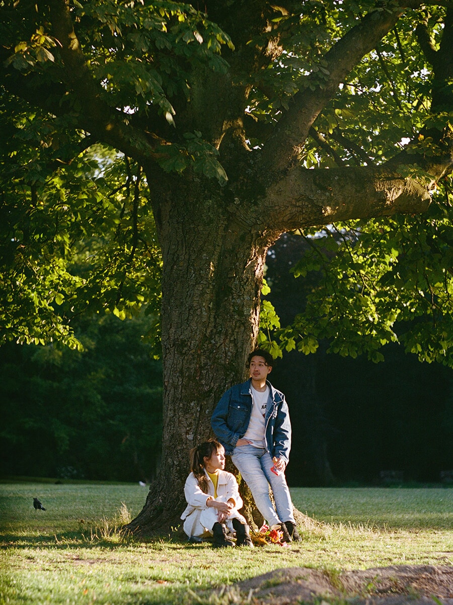 An analog couple portrait in an Amsterdam park, with a woman seated at a large tree and her partner leaning against it