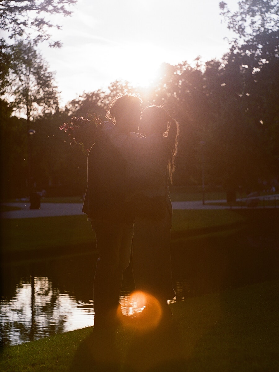 Bright backlight silhouettes a couple embracing beside a pond, with a bouquet over one shoulder and an orange flare below