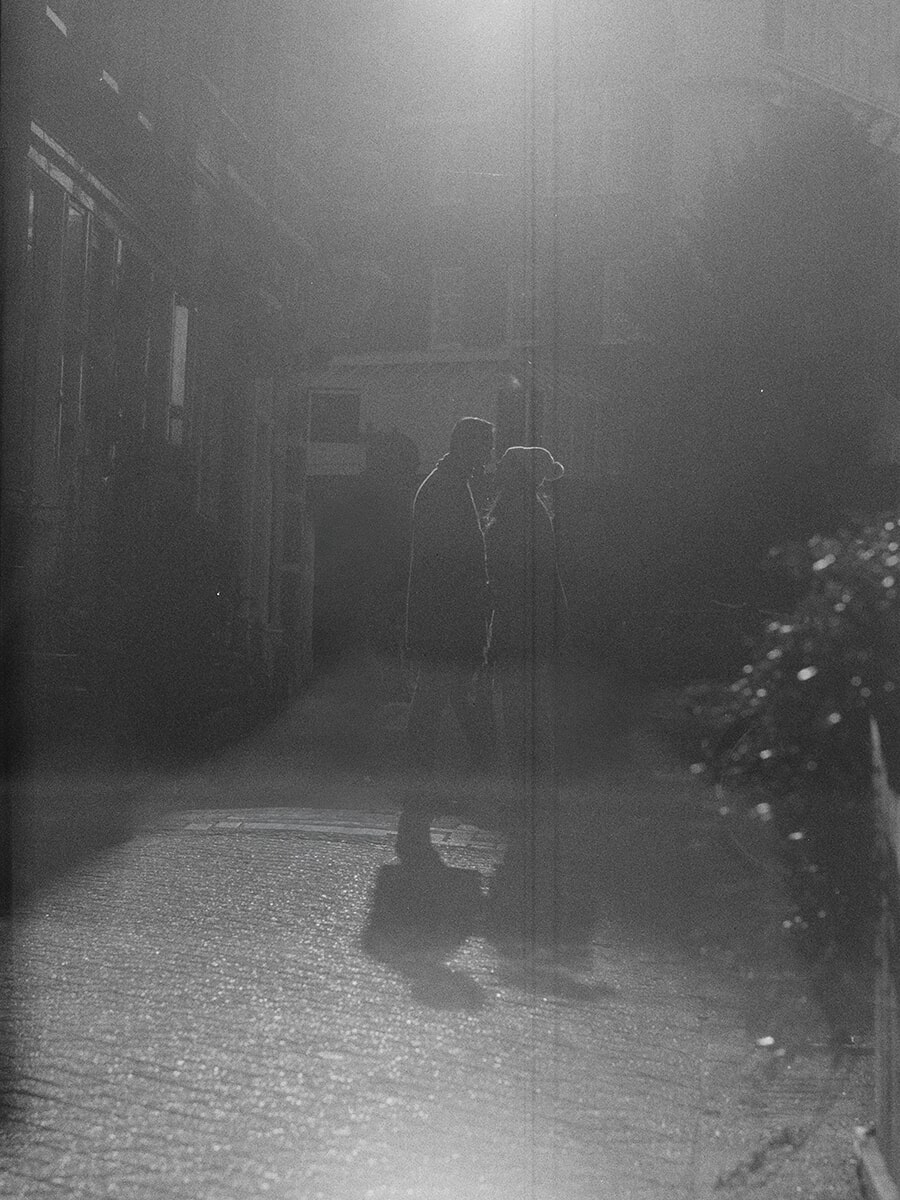 Silhouettes of a couple facing each other on a brick lane, one wearing a wide-brimmed hat, with long shadows across the paving